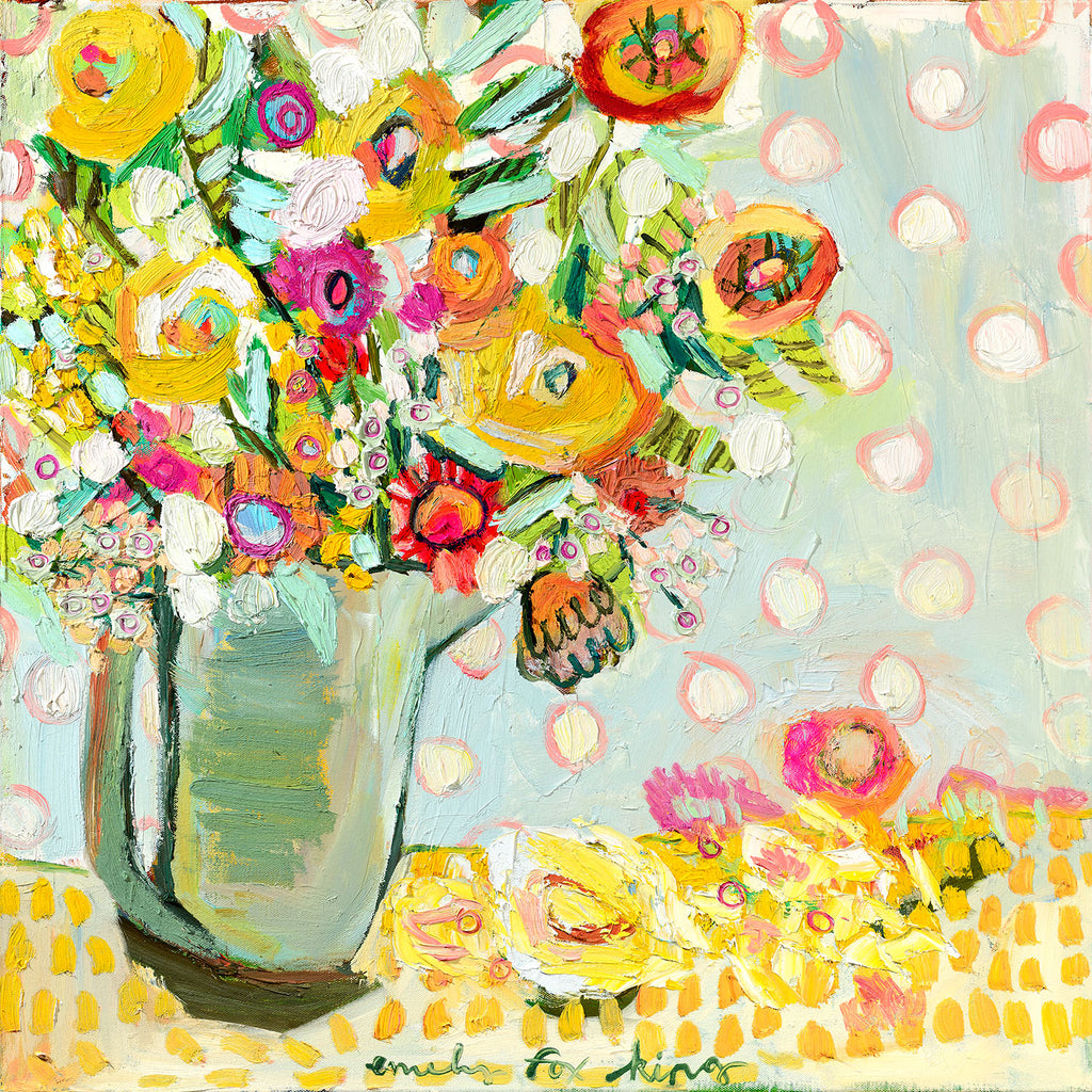 A pitcher filled with bright yellow, orange and pink flowers with green and turquoise foliage against a light blue, white and pink polka dotted background on an orange and yellow squared cloth.